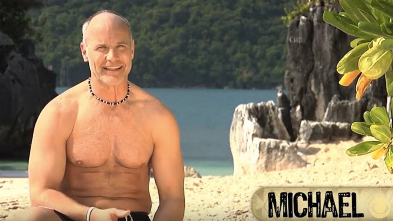 A smiling man with a shaved head and a necklace sits on a beach, wearing no shirt, with tropical plants and rocky formations in the background.