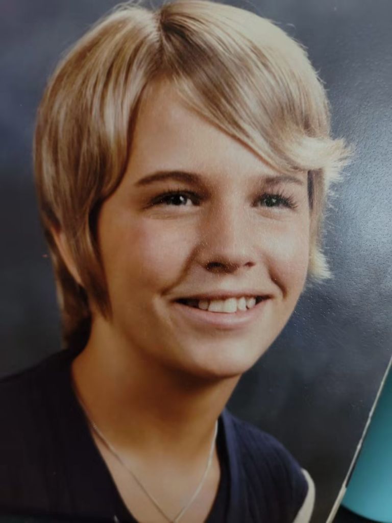 A young person with short, styled blonde hair smiling at the camera against a blurred background.