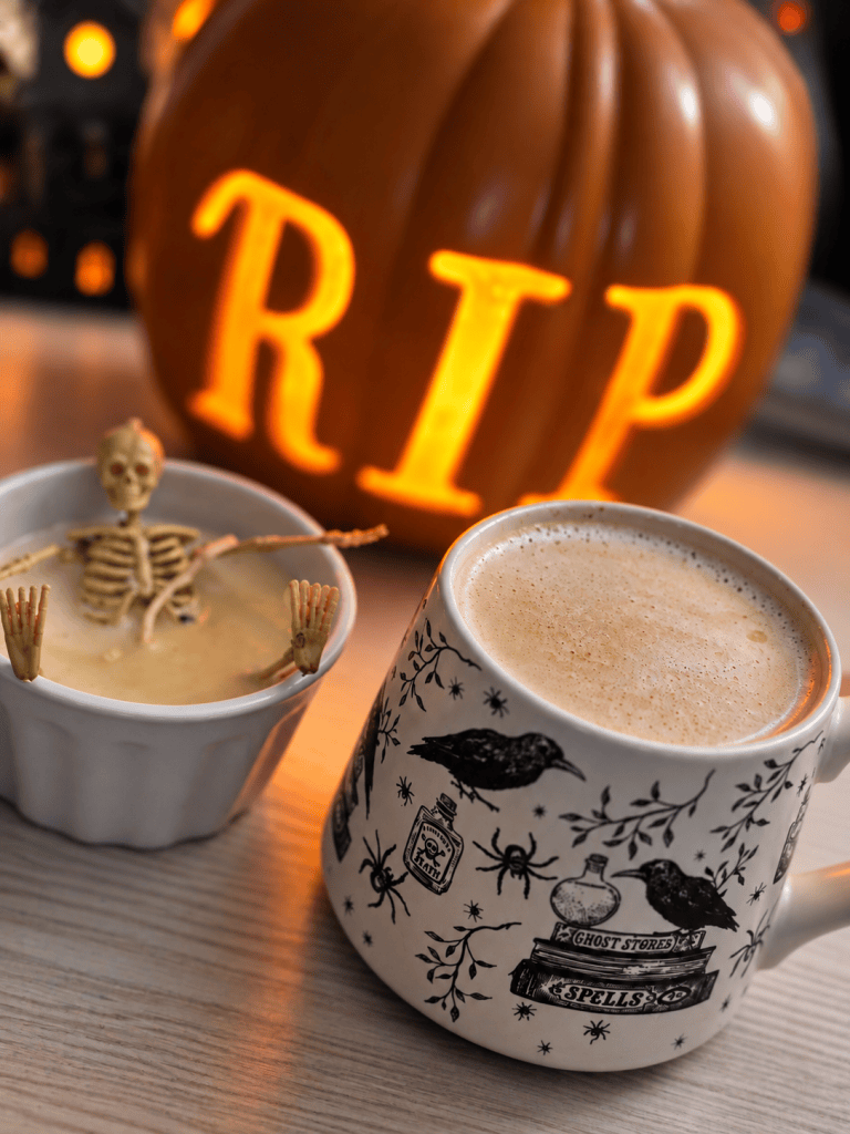 A decorated mug filled with frothy drink sits next to a small bowl containing a skeleton figure, with a carved pumpkin displaying 'RIP' in the background.